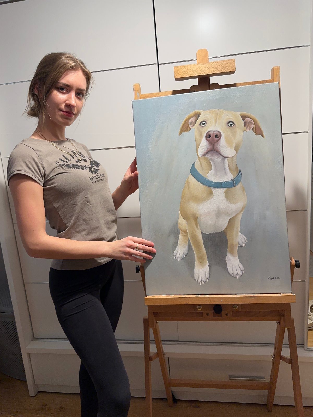 Woman standing beside a wooden easel displaying a painted portrait of a tan dog.