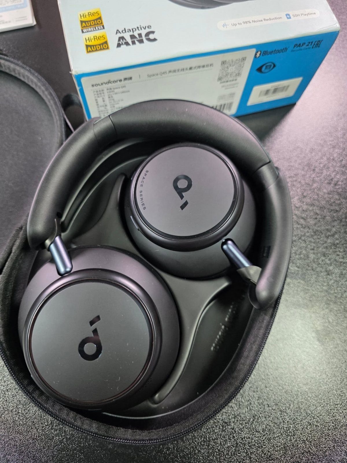 Black over-ear headphones in a carrying case with their product box behind them.