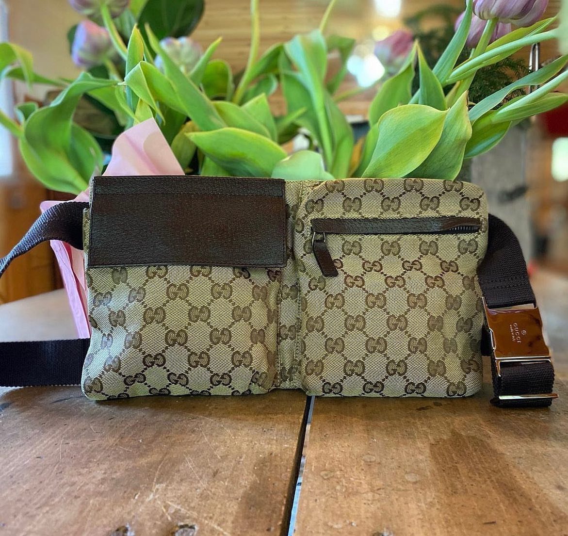 Gucci monogrammed belt bag sitting on a wooden table with flowers behind it.