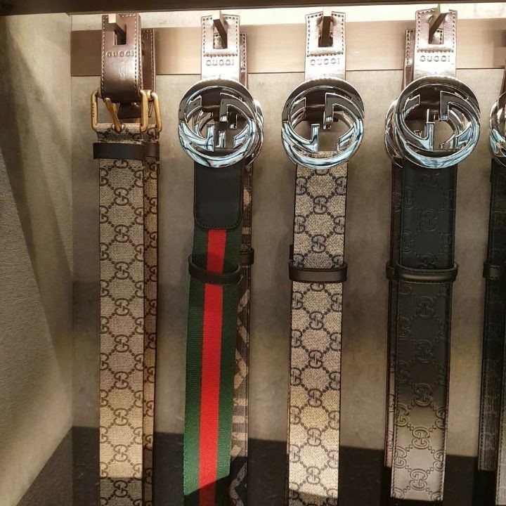 Four luxury designer belts with silver buckles hanging in a retail display.