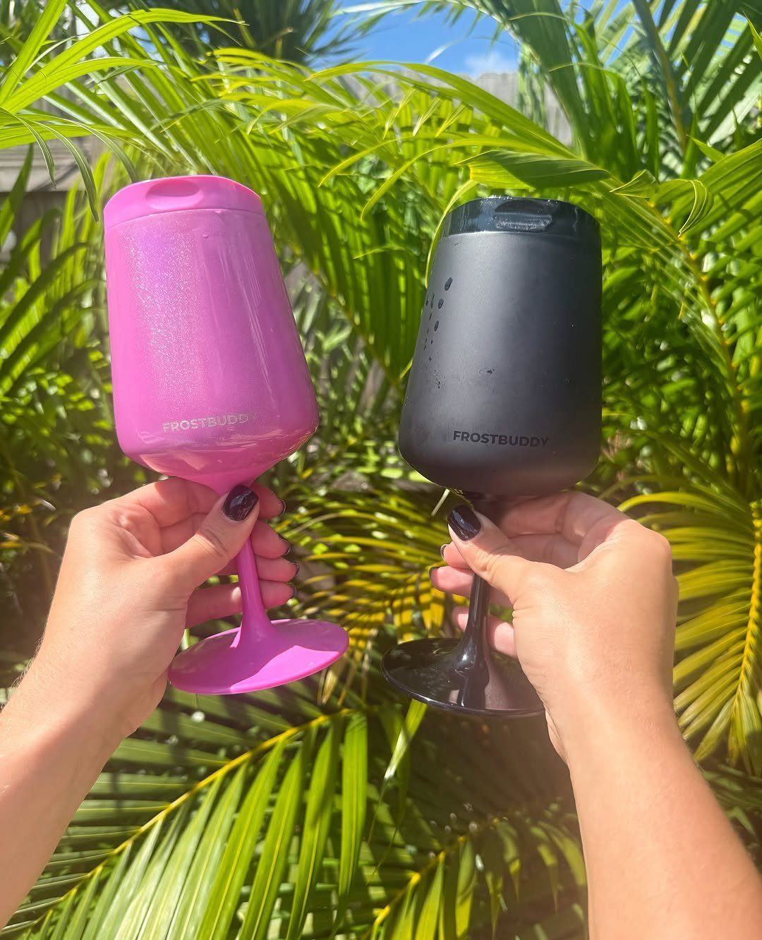 Two insulated wine tumblers, one pink and one black, held against palm leaves.
