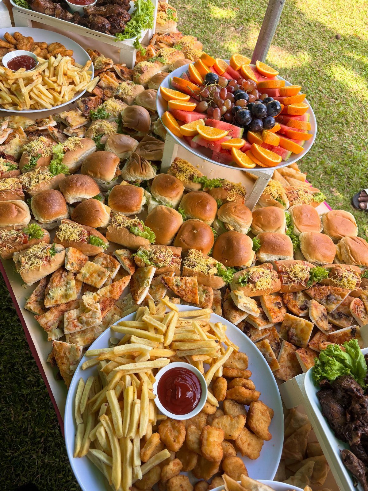Large outdoor food buffet with sliders, french fries, nuggets, and fresh fruit platters.