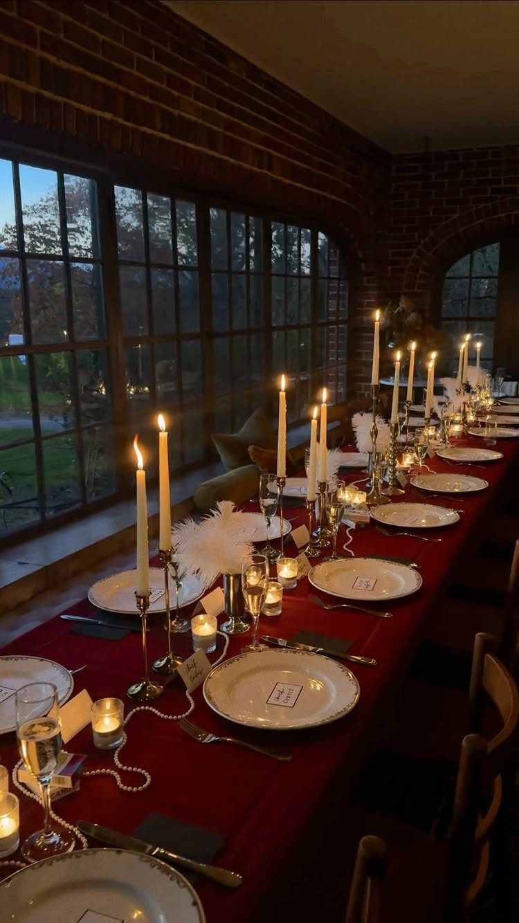 Elegant dinner table setting with red cloth, white plates, and many lit candles.