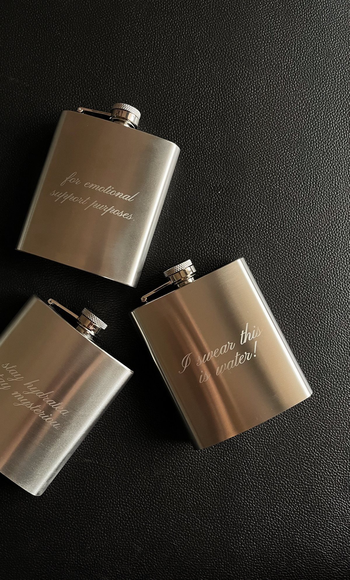 Three silver metal hip flasks with engraved cursive text on a black background