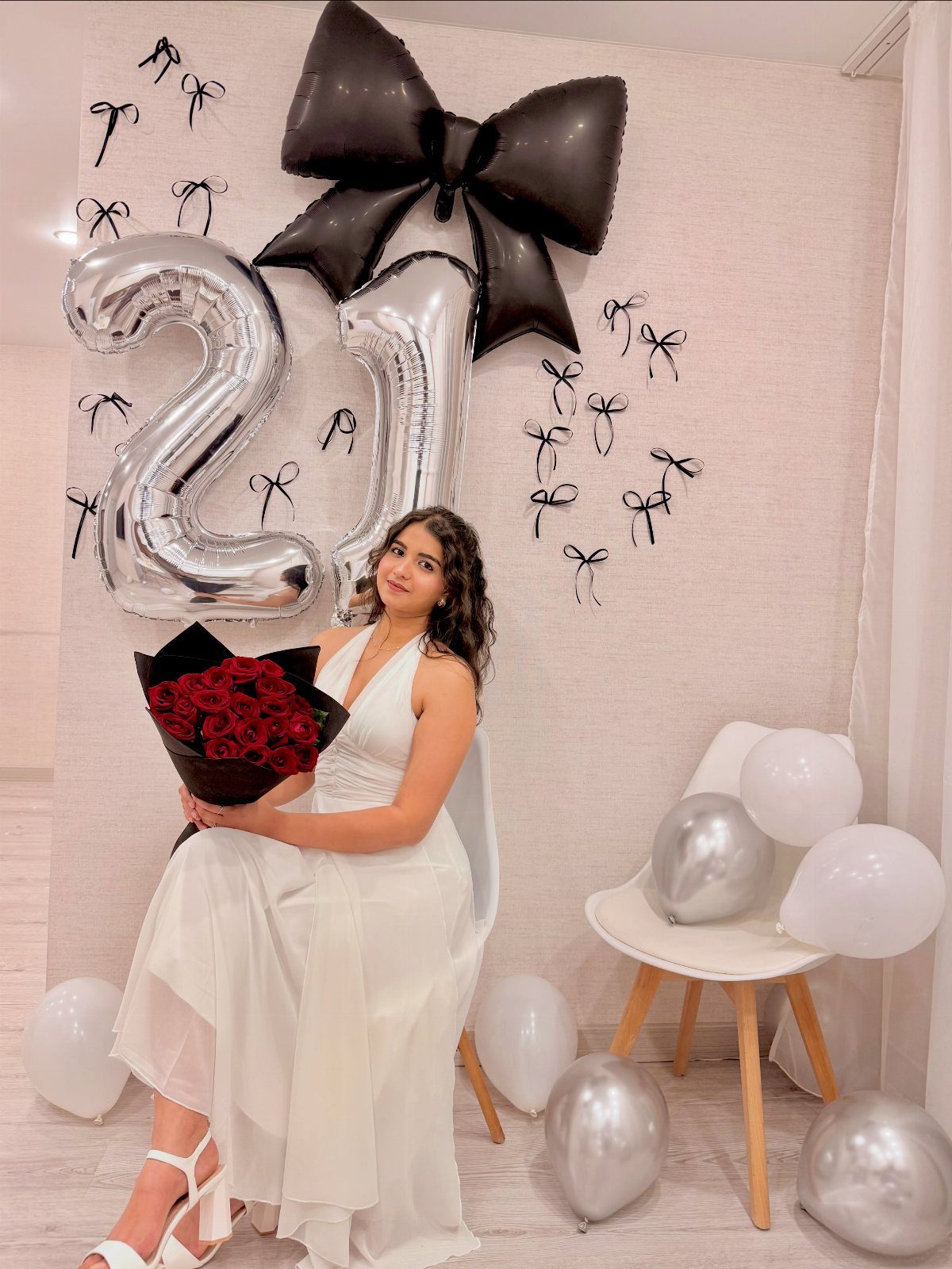 Woman in white dress posing with 21 balloons and red roses for birthday.