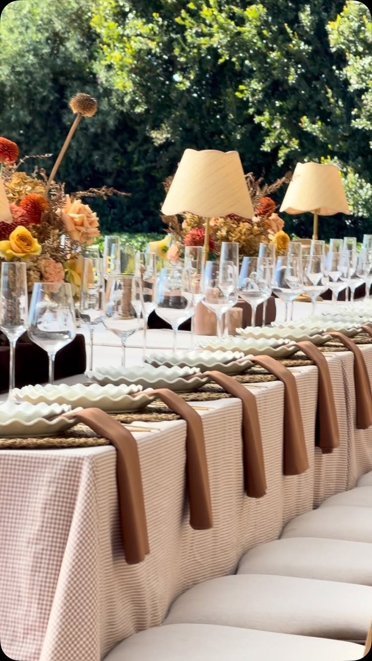 Long outdoor dining table with white checkered cloth, wine glasses, and floral centerpieces.