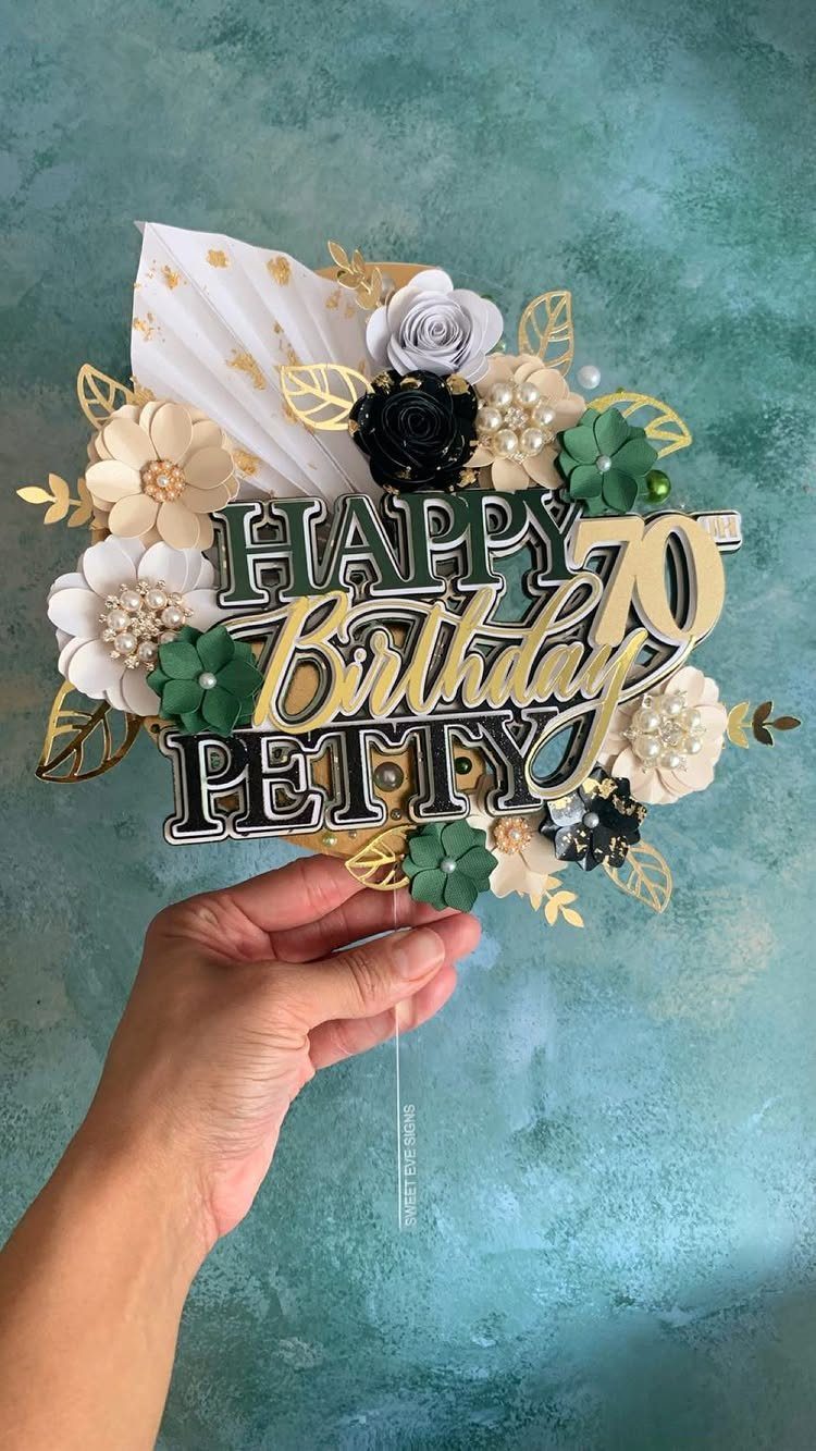 Decorative 70th birthday cake topper with paper flowers and gold leaves held by hand.