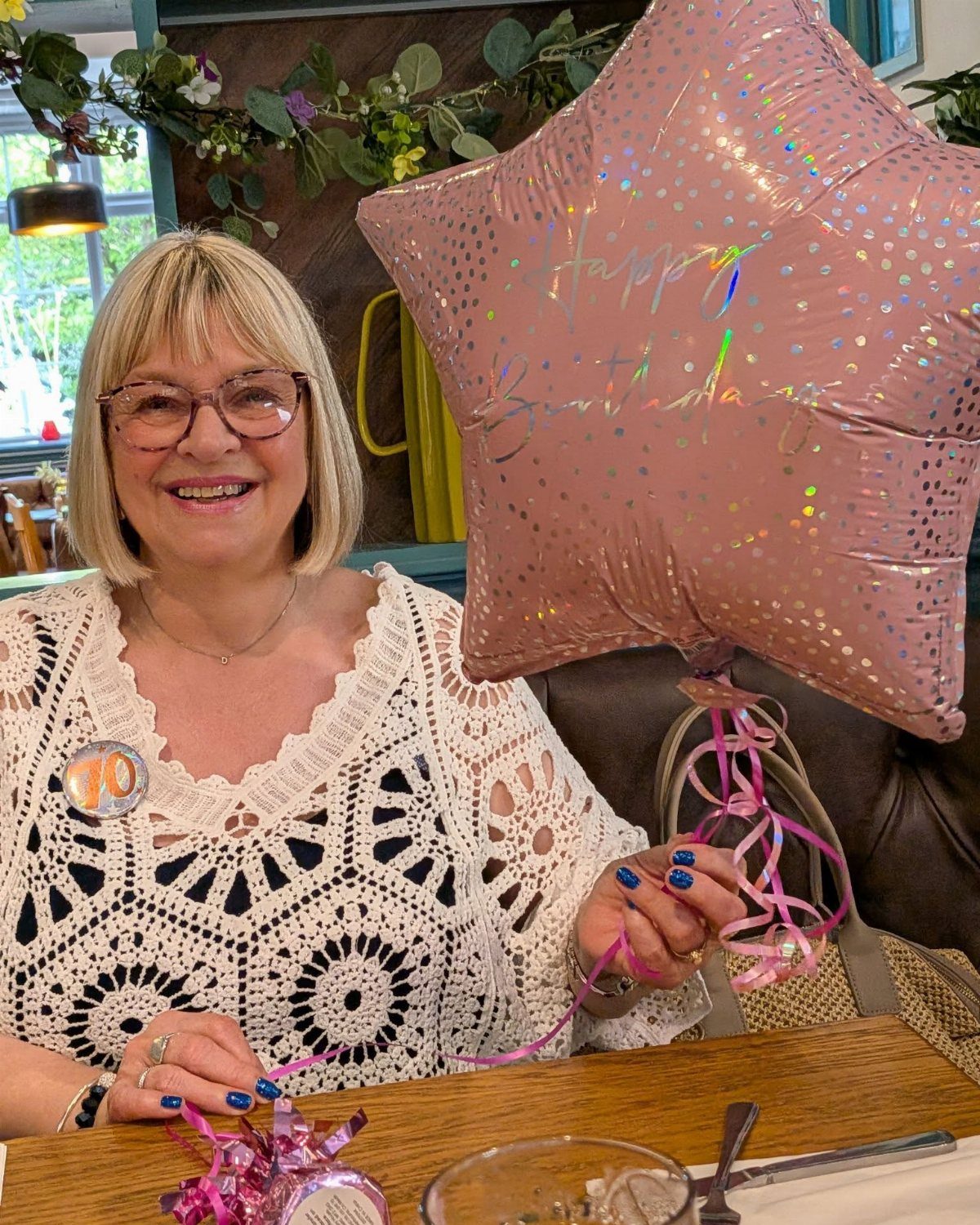 Smiling woman with blonde hair and glasses celebrating her 70th birthday with balloons.