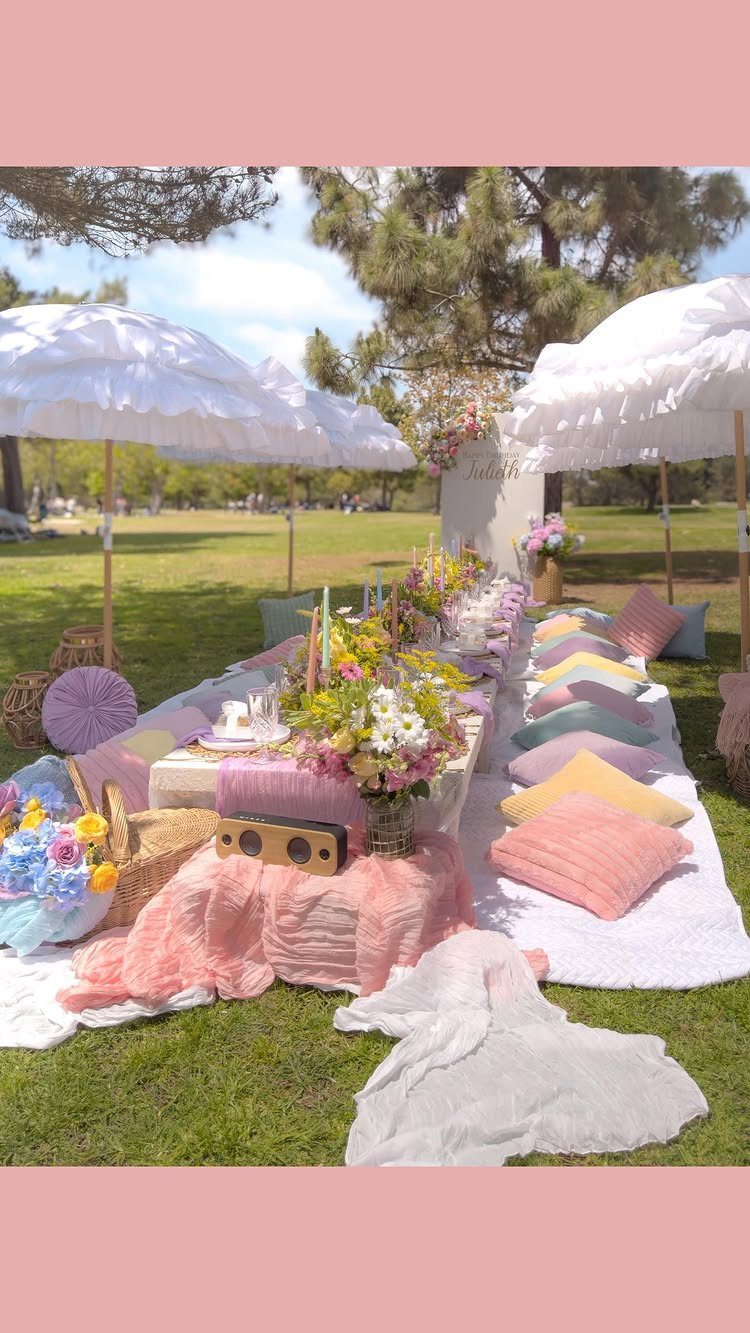 Outdoor picnic setup with white umbrellas, pink fabrics, and colorful cushions on grass.