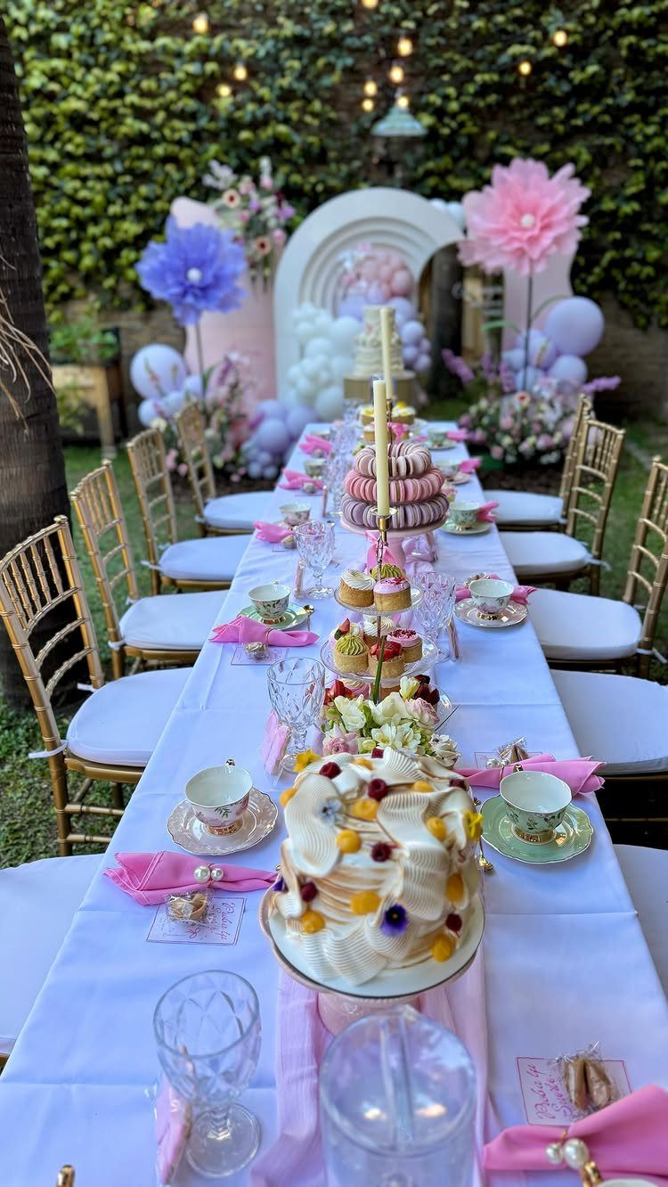Elegant outdoor birthday table setting with cakes, pink decorations, and floral balloon arch.