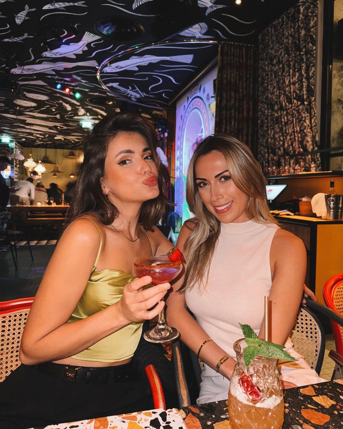 Two women posing with cocktails in a dimly lit restaurant with fish-patterned ceilings.