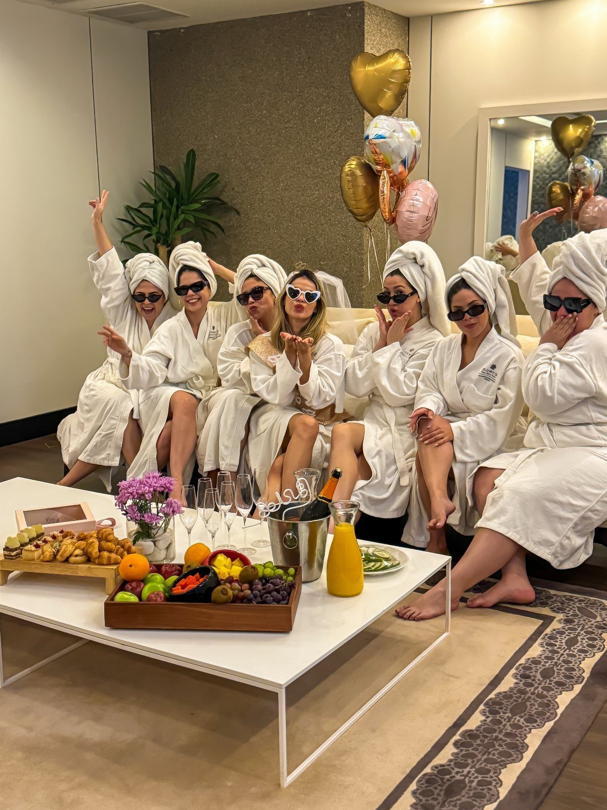 Seven women in white spa robes and towels posing with champagne and fruit.