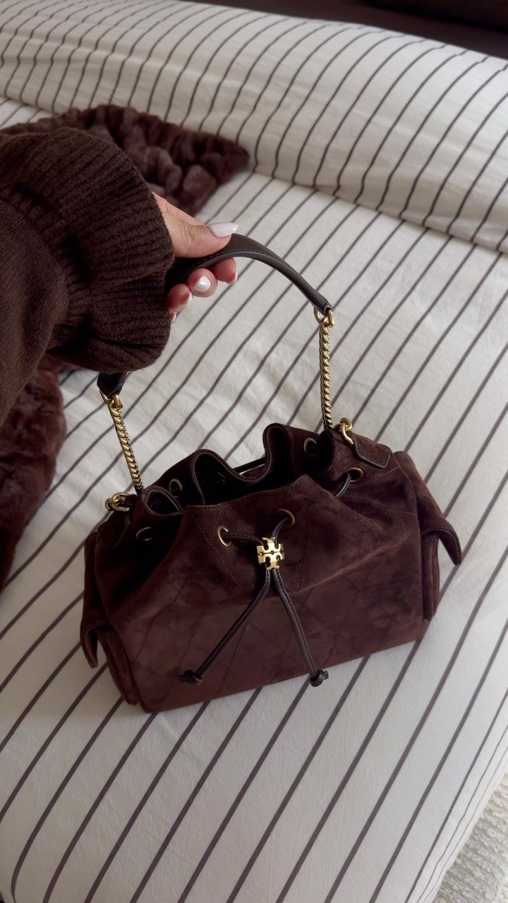 Brown suede luxury drawstring handbag with gold chain handles on striped fabric.