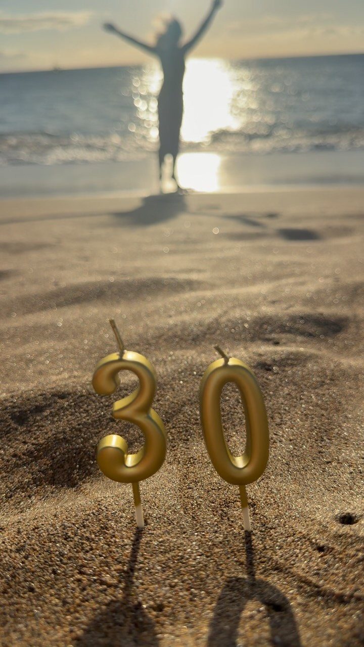 Gold number 30 candles in beach sand with a silhouette at sunset.