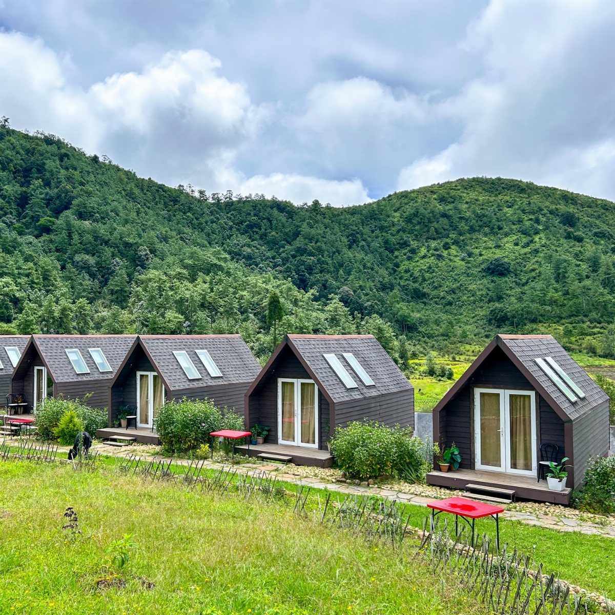 A-frame cabins on a grassy hillside with green mountains under a cloudy sky.