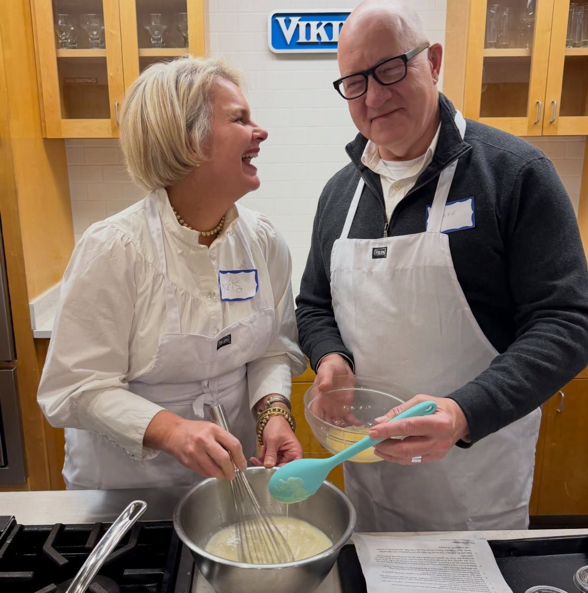 Older couple in white aprons cooking together in a kitchen with a stove.