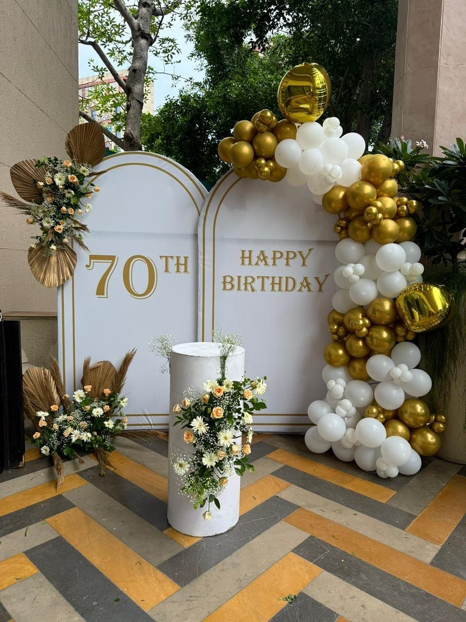 White and gold 70th birthday backdrop with balloon arch and floral arrangements outdoors