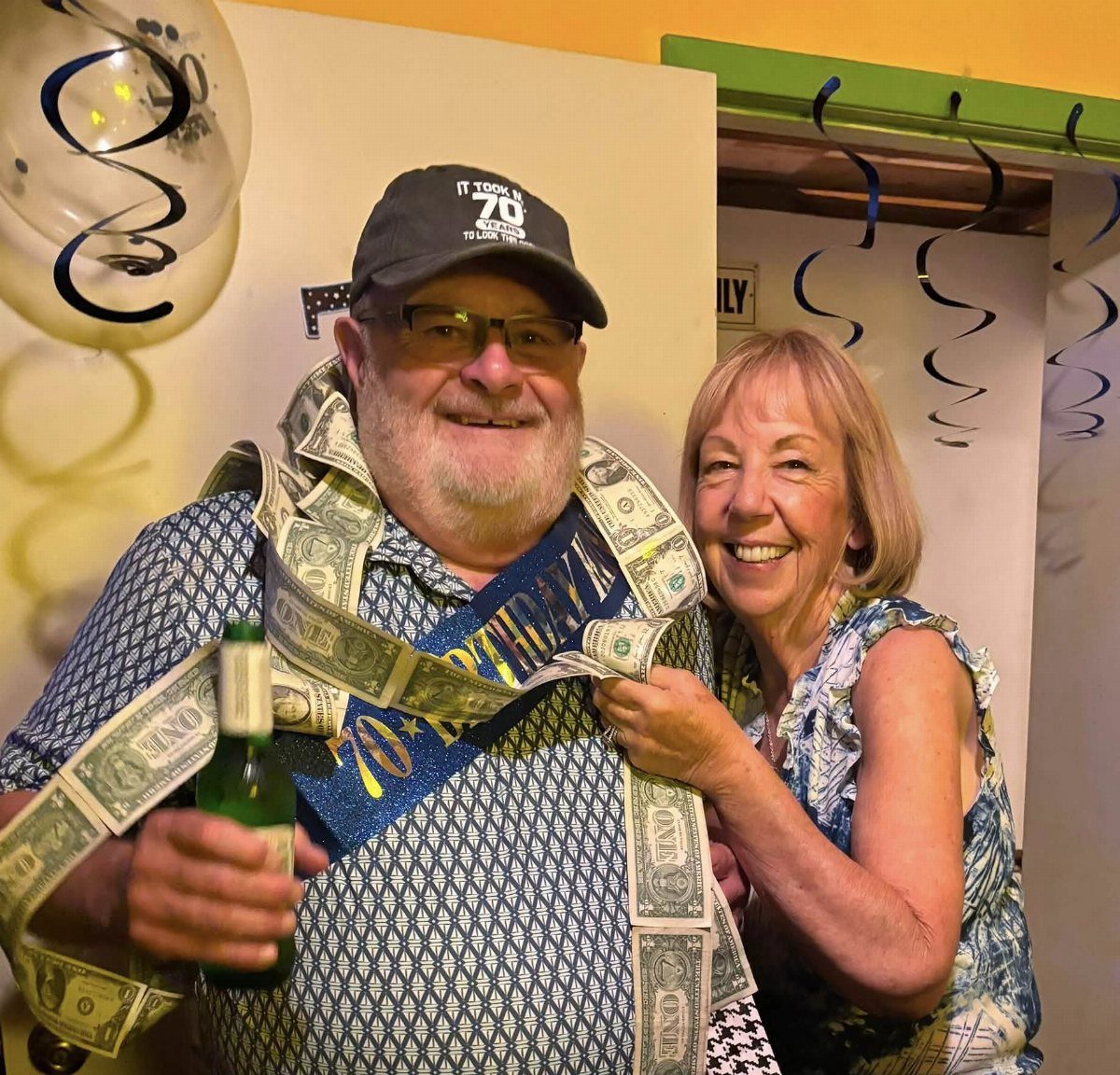 Older man wearing a 70th birthday cap and dollar bill sash with a woman.