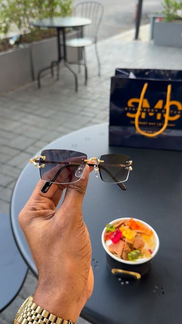 Gold-rimmed sunglasses held over a black table with a shopping bag and food.