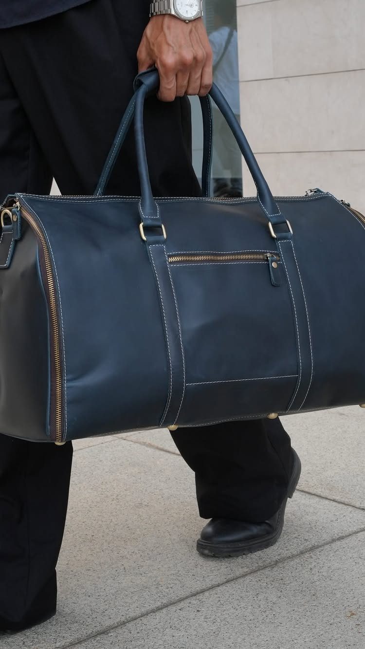 Navy blue leather duffel bag held by a man in black trousers.