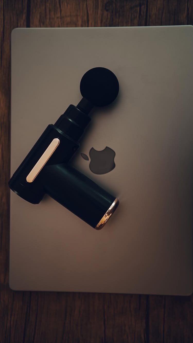Black massage gun with gold accents resting on a grey laptop lid.