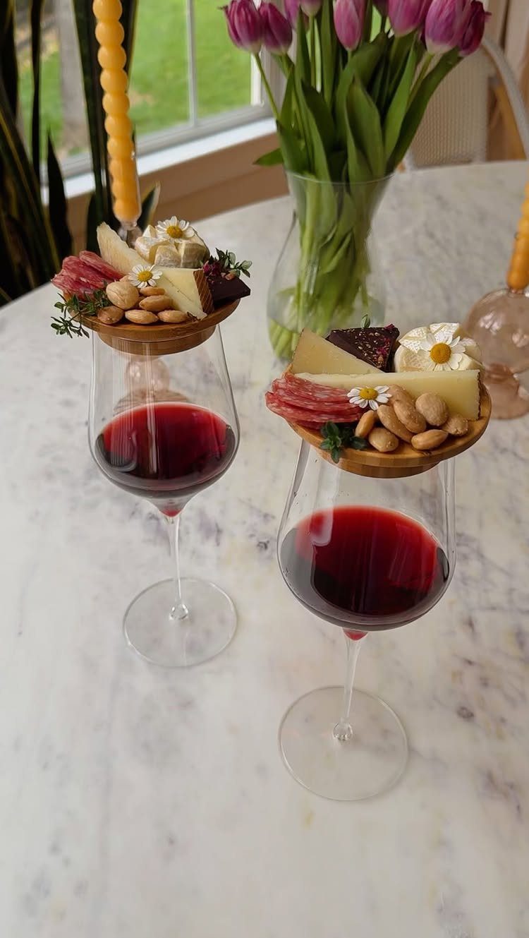 Two red wine glasses with wooden charcuterie platters balanced on the rims.