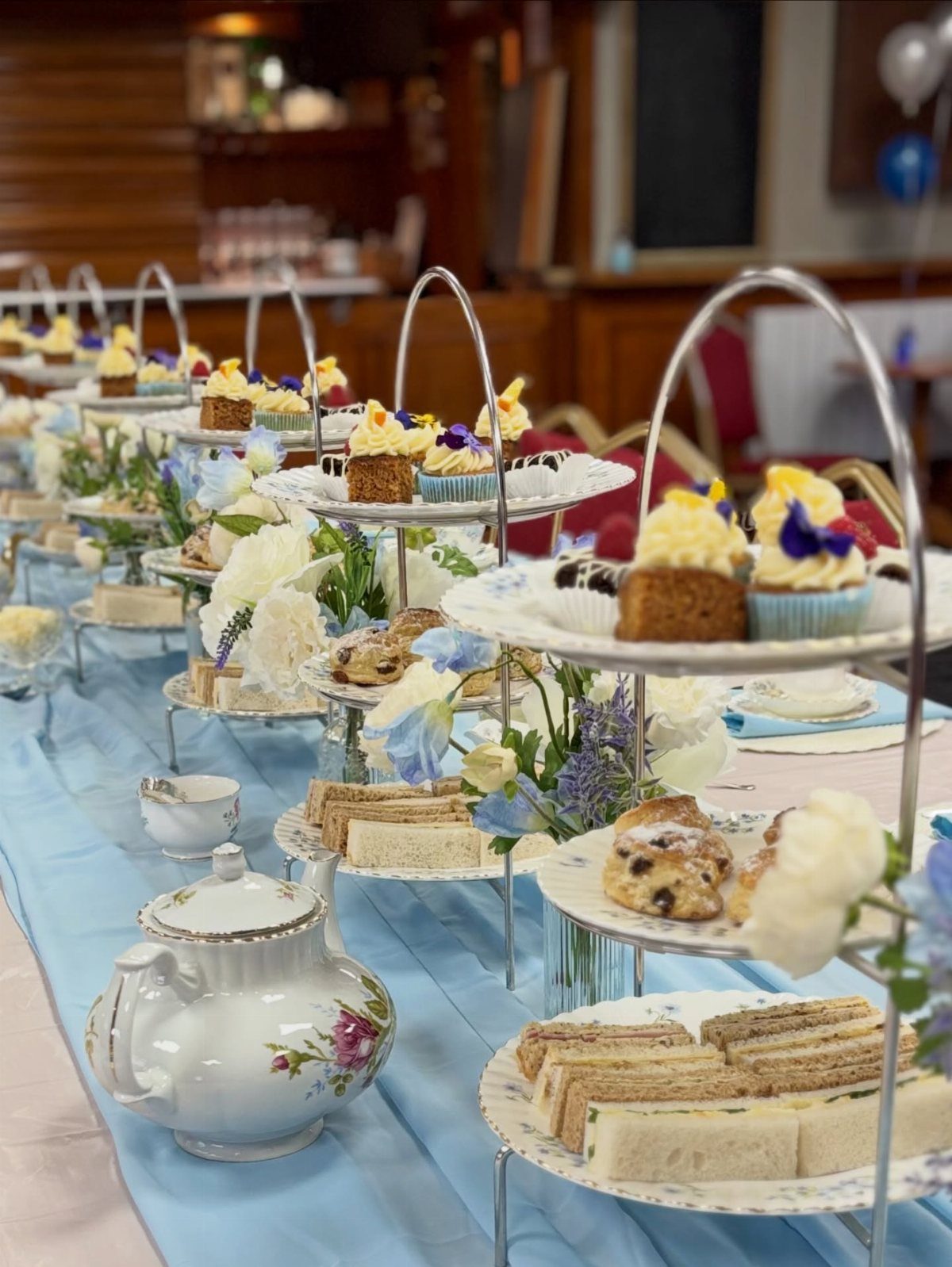 Tiered cake stands with sandwiches and cupcakes on a blue tablecloth with a teapot.