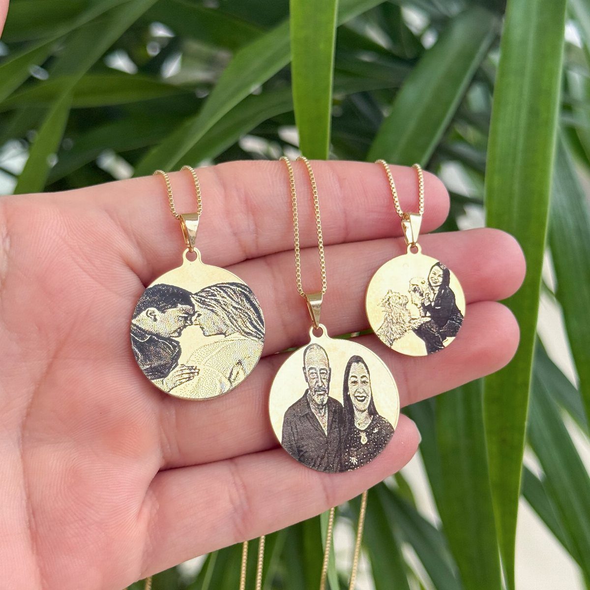 Three gold circular pendant necklaces with engraved photos held against green leaves.
