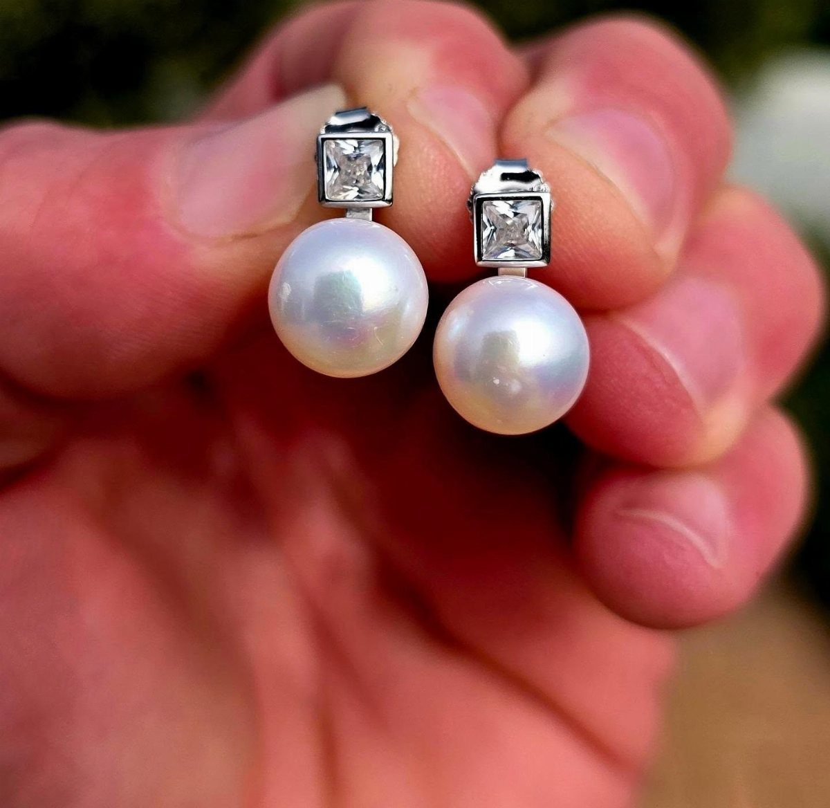 Pearl earrings with square gemstones held in a person's hand against a blurred background.
