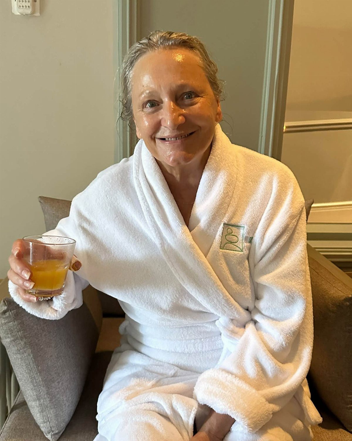 Older woman in a white bathrobe smiling and holding a glass of orange juice.