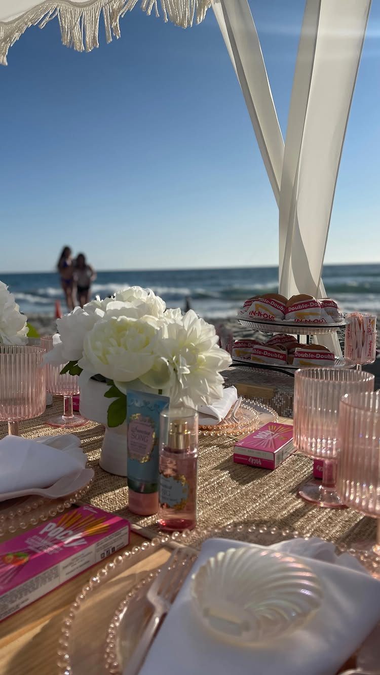 Beach picnic table with white flowers, pink glassware, and treats under a white canopy.