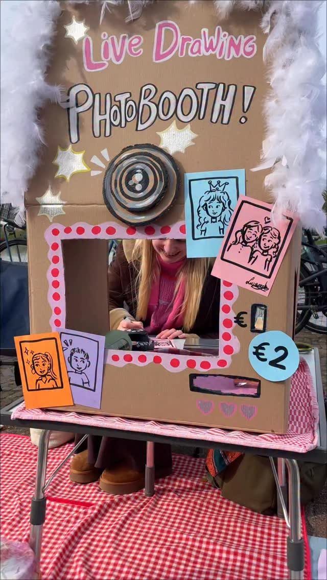 Woman drawing inside a cardboard camera booth labeled Live Drawing PhotoBooth