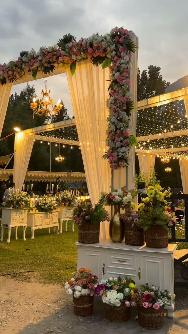 Outdoor event decoration with white drapes, floral arches, and gold chandeliers on grass.