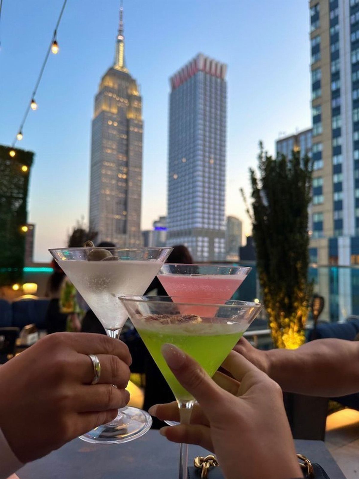 Three colorful cocktails being toasted on a rooftop with New York City skyline background