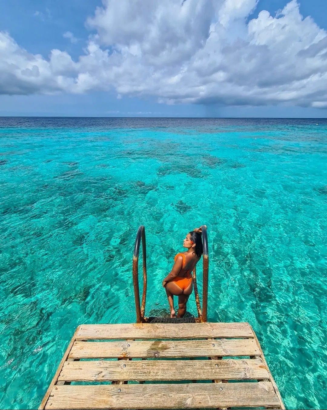 Woman in orange bikini standing on a pier ladder over turquoise ocean water