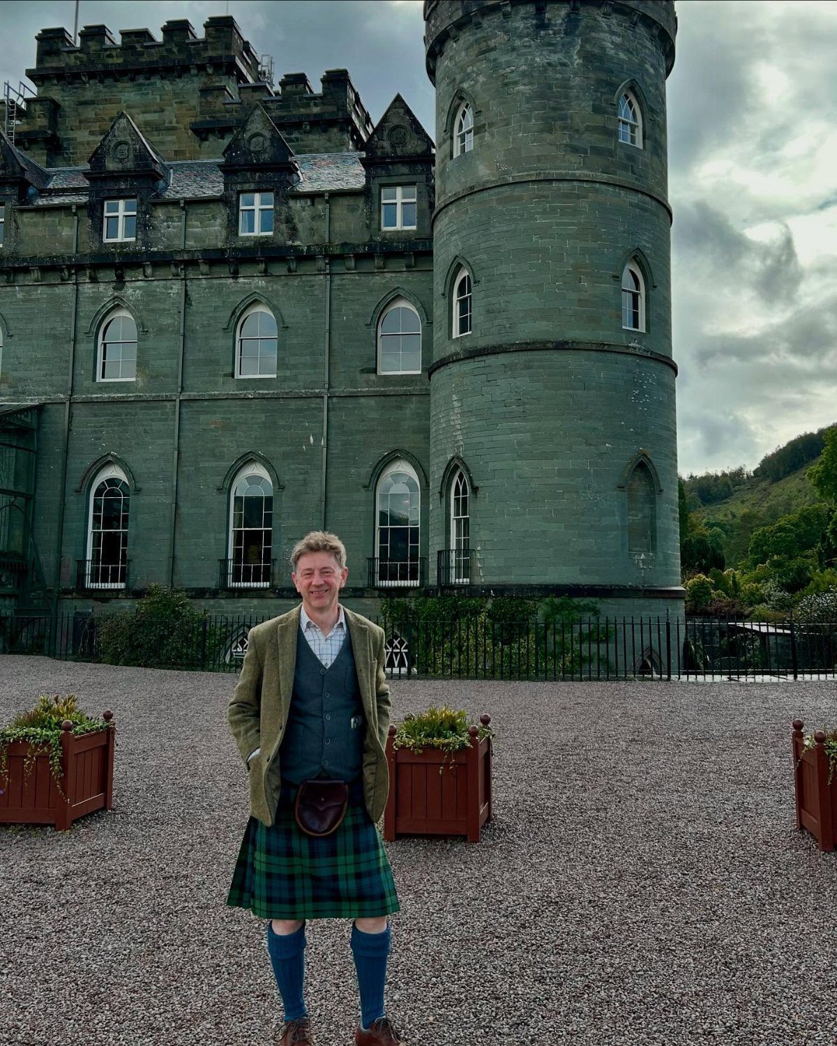 Man in traditional Scottish kilt standing before a large grey stone castle tower.