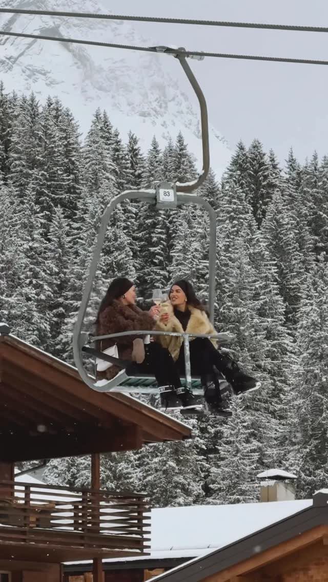 Two women drinking champagne on a chairlift amidst snowy mountains and evergreen trees.