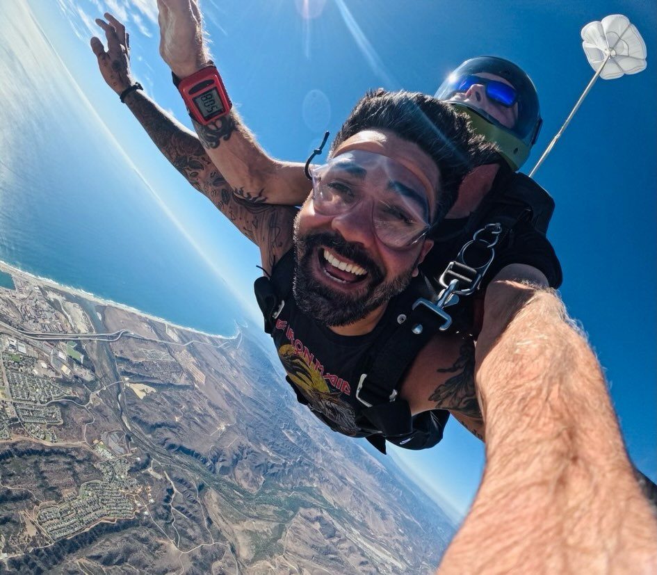 Smiling man taking a selfie while skydiving tandem over a coastal landscape