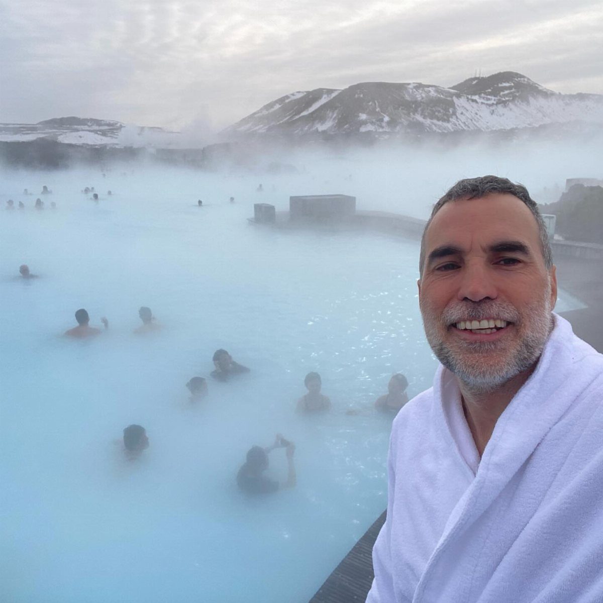 Smiling man in white bathrobe at a steaming blue geothermal spa with mountains.
