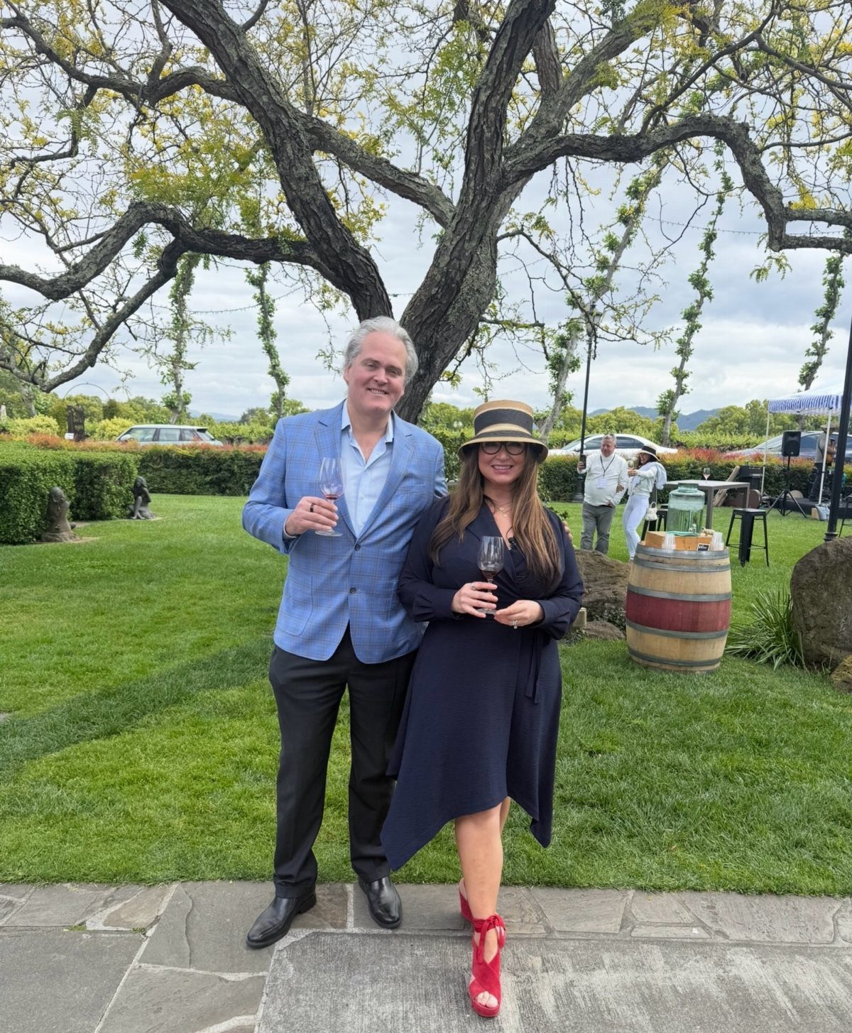 Couple holding wine glasses standing on a vineyard lawn next to a barrel.