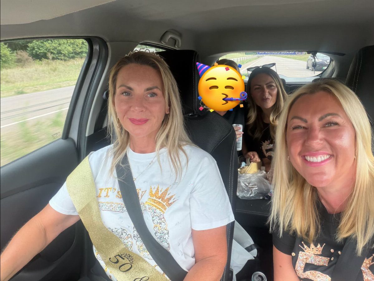 Four women smiling inside a car, one wearing a gold 50th birthday sash.