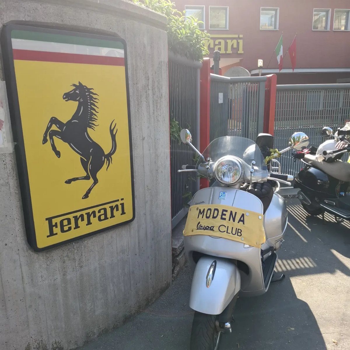 Silver Vespa scooter parked next to a Ferrari logo sign in Modena, Italy.