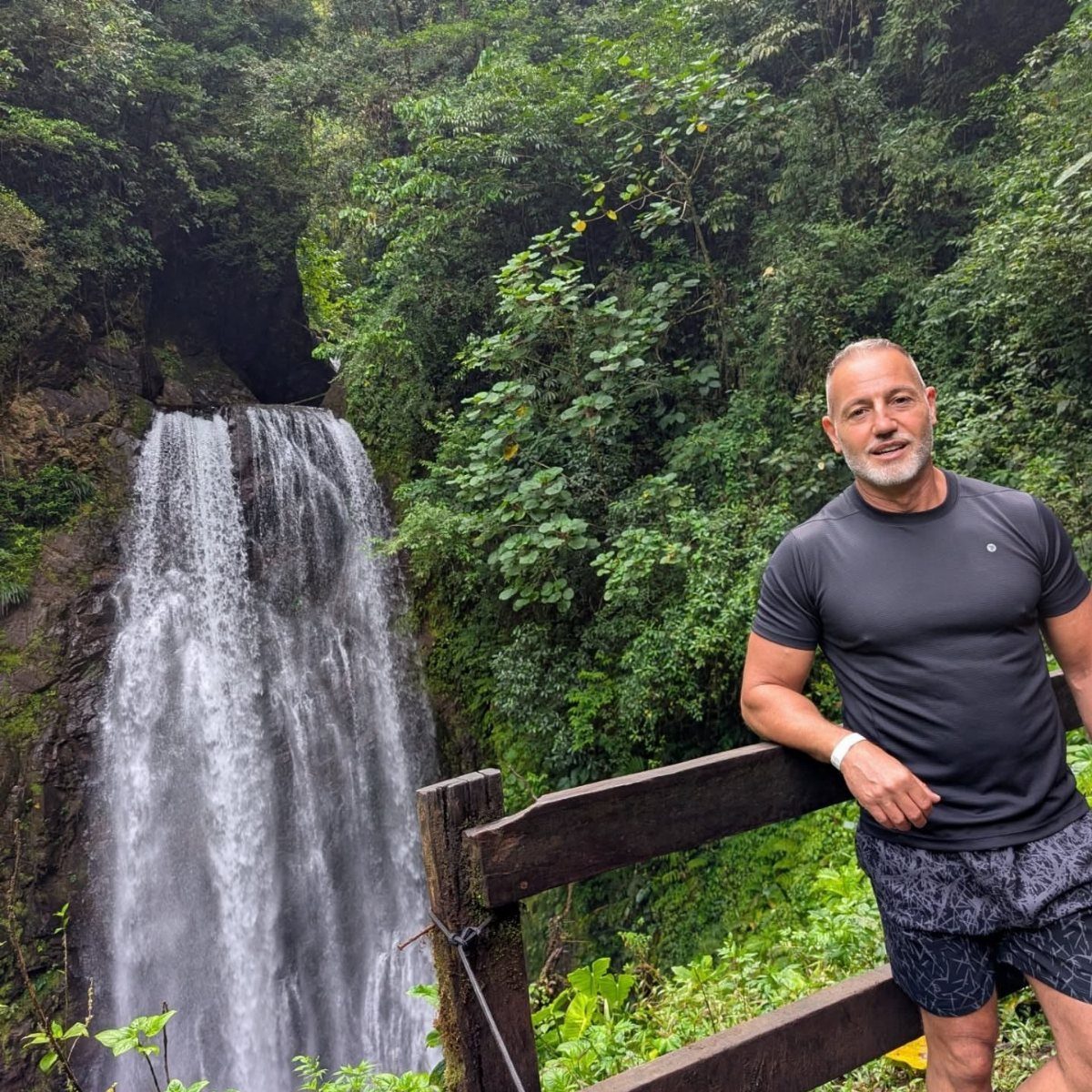 Man leaning on a wooden railing with a tropical waterfall and lush greenery behind.