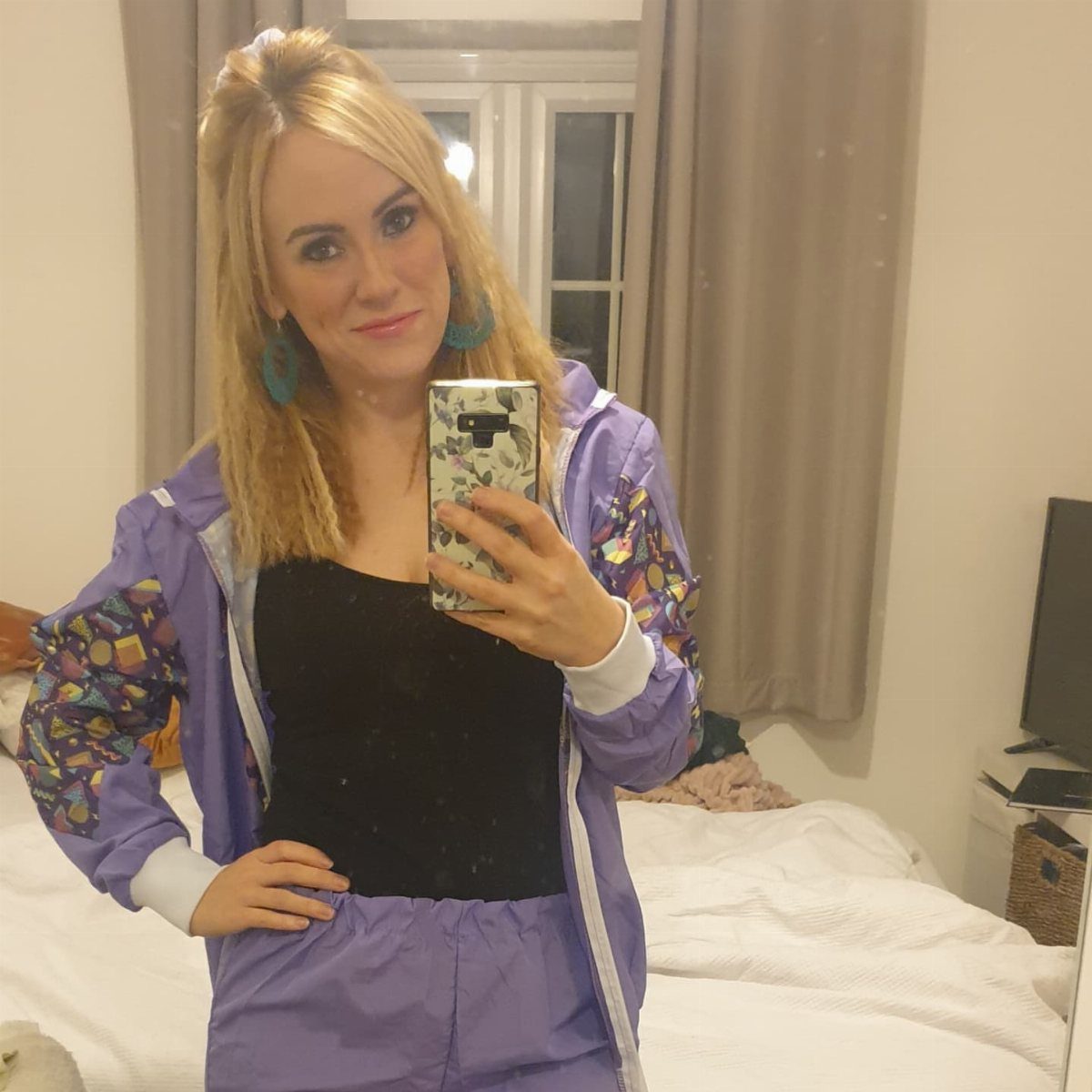 Woman in purple 1980s tracksuit and blue hoop earrings taking a mirror selfie.