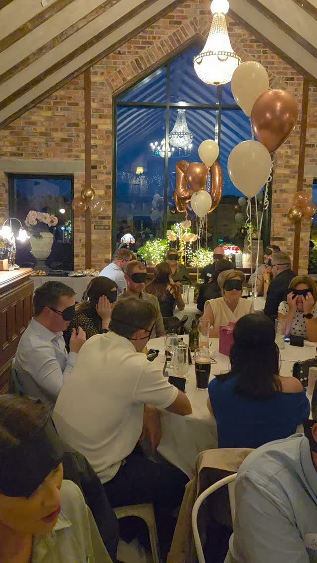 Guests wearing blindfolds at a 40th birthday party with gold and white balloons.