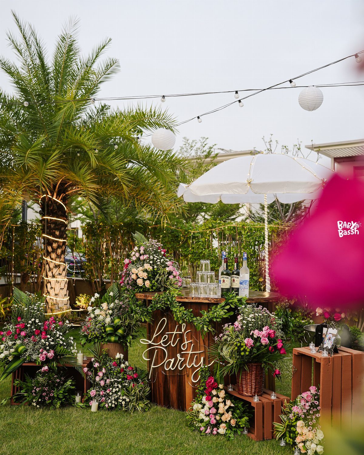 Outdoor party bar decorated with pink flowers, a neon Let's Party sign, and string lights.
