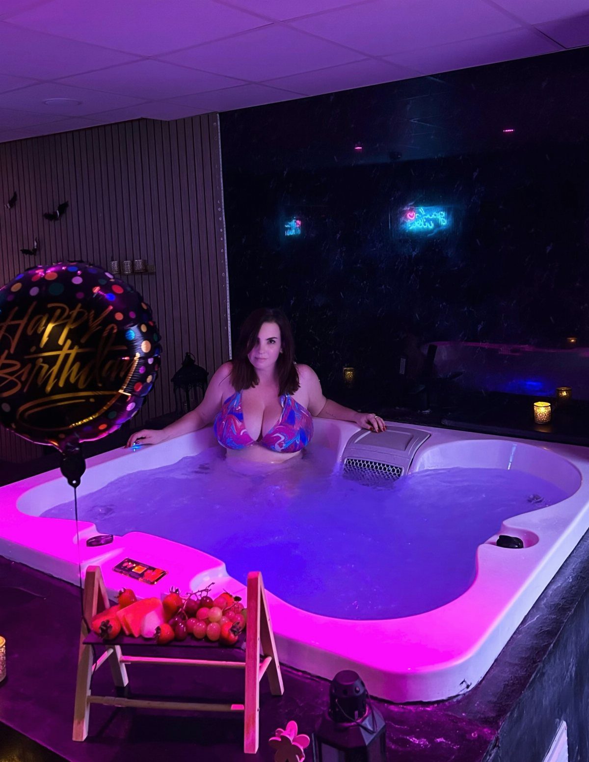 Woman in a bikini relaxing in a purple glowing hot tub with birthday balloon.