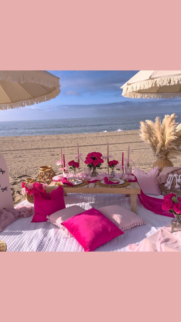 Beach picnic setup with pink pillows, flowers, and umbrellas on the sand by the ocean.