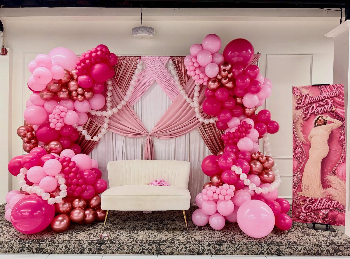 Pink and rose gold balloon arches framing a white loveseat and draped backdrop.