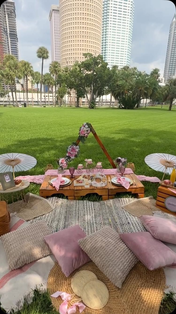 Luxury picnic setup with low wooden table, pink cushions, and floral teepee on grass.